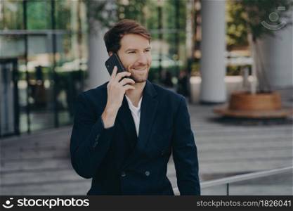 Confident man entrepreneur dressed in formal clothes makes phone call in roaming looks away has cheerful expression poses near office building enjoys telephone conversation during work break. Confident man entrepreneur dressed in formal clothes makes phone call in roaming