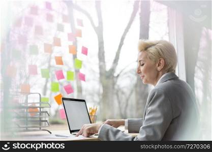 Confident mature businesswoman using laptop while sitting with document at desk in office
