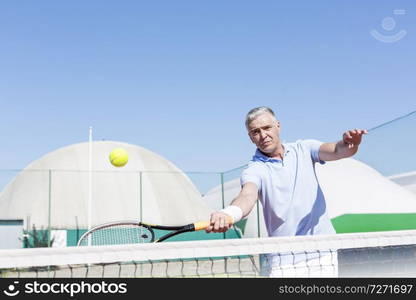 Confident mature man hitting tennis ball with racket on court against clear blue sky