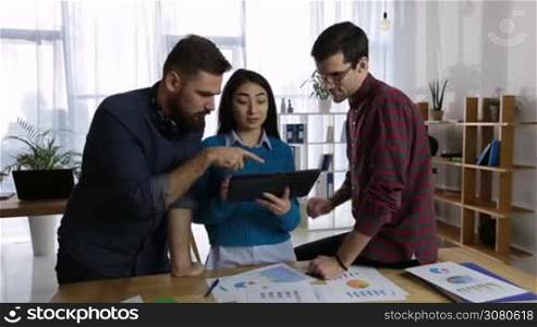 Confident multi-ethnic business team working together and using digital tablet to achieve better results in modern office. Friendly hipster colleagues with touchpad working with financial data. Dolly