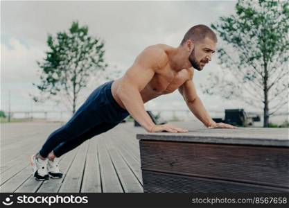 Confident muscled sportsman in sport wear doing push up outdoors, works on biceps and trains determination. Strong topless athletic man exercises in open air. People, motivation, training concept