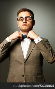 Confident nerd in eyeglasses adjusting his bow-tie against grey background