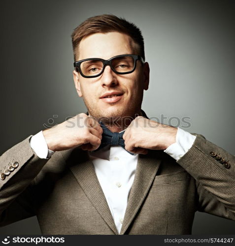 Confident nerd in eyeglasses adjusting his bow-tie against grey background