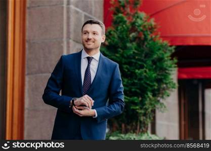 Confident pleased businness worker poses outdoor, wears elegant suit and wrist watch, has happy look into distance, notices his business partner, glad to cooperate and achieve great results.