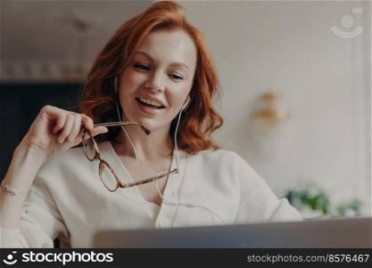 Confident pleased redhead woman concentrated in laptop computer, watches video or movie on laptop computer, enjoys friendly video call, holds transparent glasses, uses free internet connection