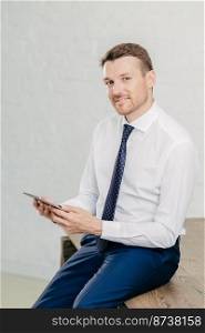 Confident pleased young male entrepreneur in luxury formal clothes, reads recieved text message from colleague on modern tablet computer, looks joyfully at camera, poses in office. Vertical shot