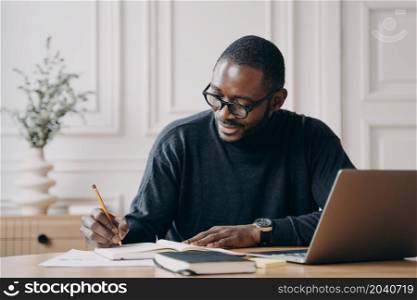 Confident positive afro american businessman working remotely online while sitting at home office, getting ready for monthly financial report, making notes in agenda while checking charts on laptop. Confident young Afro american businessman working remotely online while sitting at home office