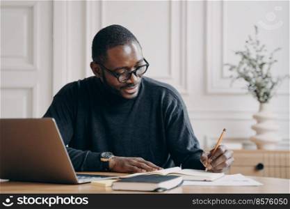 Confident positive afro american businessman working remotely online while sitting at home office, getting ready for monthly financial report, making notes in agenda while checking charts on laptop. Confident young Afro american businessman working remotely online while sitting at home office
