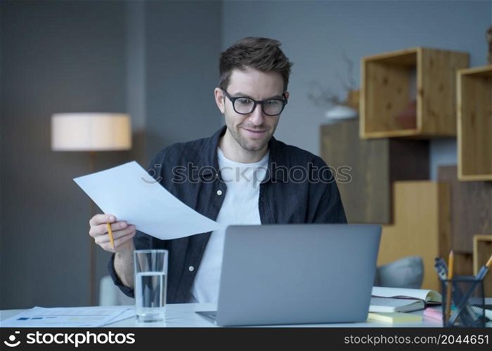 Confident positive man financial counselor in eyewear works remotely online at modern home office. Smiling company employee holding paper documents, chatting online with clients on laptop at workplace. Confident positive man financial counselor in eyewear works remotely online at modern home office