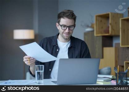 Confident positive man financial counselor in eyewear works remotely online at modern home office. Smiling company employee holding paper documents, chatting online with clients on laptop at workplace. Confident positive man financial counselor in eyewear works remotely online at modern home office