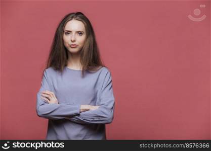 Confident serious attractive female model keeps hands folded, wears casual clothing, listens attentively information, poses against pink background with blank copy space for your promotional text