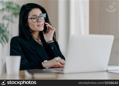 Confident serious business lady has phone conversation, poses in front of laptop computer, wears optical glasses for vision correction, poses at work place, makes reserach work, dressed formally