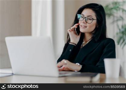 Confident serious business lady has phone conversation, poses in front of laptop computer, wears optical glasses for vision correction, poses at work place, makes reserach work, dressed formally