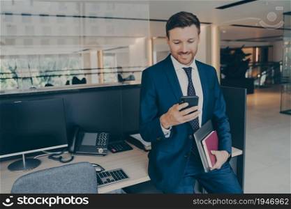 Confident smiling entrepreneur with laptop and agenda using smartphone and smiling, reading emails, checking last financial reports, while waiting for assistant to start business presentation. Confident smiling entrepreneur using smartphone and smiling while standing in office