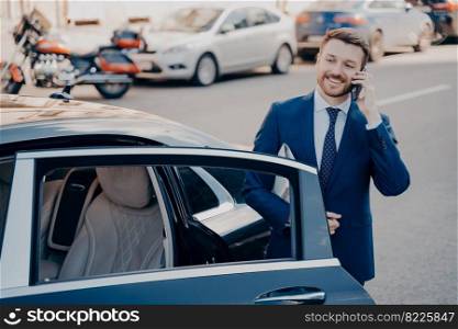 Confident smiling executive dressed in blue formal suit leaving black limousine after arriving at his workplace, talking on phone, holding newspaper while standing alone next to car with open door. Confident executive talking on phone next to limousine