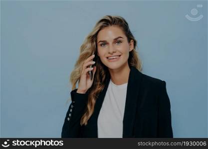 Confident smiling young office worker talking on mobile phone, looking straight at camera while standing isolated over blue background, wearing stylish black blazer over white shirt. Confident positive smiling young female office worker talking on smartphone