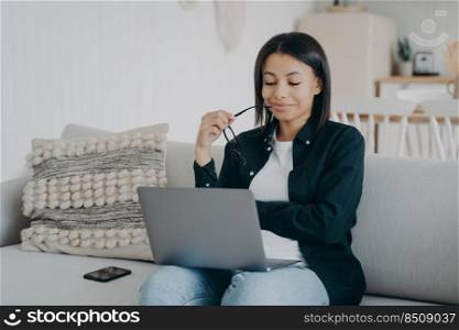Confident sophisticated arab woman at work, career concept. Businesswoman is working from home. Lady is sitting in comfortable couch with laptop and smiling, looking at the screen.. Confident sophisticated woman at work, career concept. Businesswoman is working on laptop from home.
