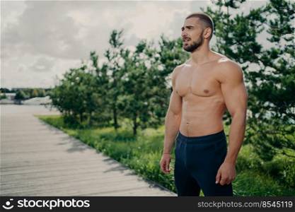 Confident sporty man bodybuilder has workout outdoor, looks thoughtfully into distance, wears shorts, has muscular body, strong biceps, stands over trees and sky background, shows strong torso