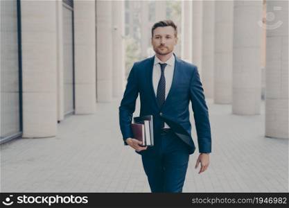 Confident successful businessman in formal clothes with laptop, notebook and book in hand walking on city street with urban background, hurrying up at business meeting with partner or client. Confident successful businessman with laptop in hand walking on city street with urban background