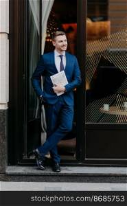 Confident successful businessman in formal suit, keeps hand in pocket, holds business magazine, stands near entrance of cozy cafeteria, has pleased expression. People, job and occupation concept