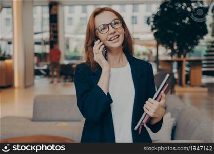 Confident successful ginger female entrepreneur holding laptop and notebook in hand, enjoying pleasant conversation on mobile phone with client, wearing eyeglasses and formal clothes. Confident successful ginger female entrepreneur enjoying pleasant conversation on mobile phone