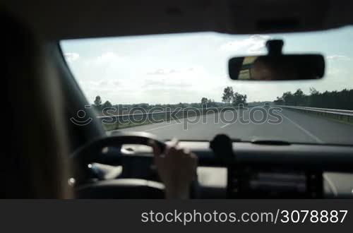 Confident woman driving car on motorway during summer road trip with beautiful landscape on background. View from inside out of vehicle&acute;s interior.