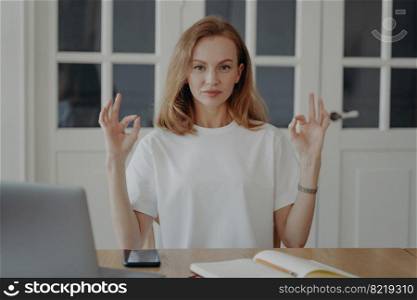 Confident woman gestures ok sitting at the desk in her apartment. Successful mid adult businesswoman. Lovely european lady in white t-shirt is entrepreneur having remote work at home office.. Confident woman gestures ok sitting at the desk in apartment. Successful mid adult businesswoman.