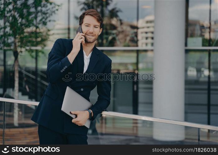 Confident young bearded businessman dressed in formal clothes making business call, standing outside of glass building with laptop in relaxed pose while talking on mobile phone with colleagues. Confident young businessman standing outside with laptop computer and talking on mobile phone