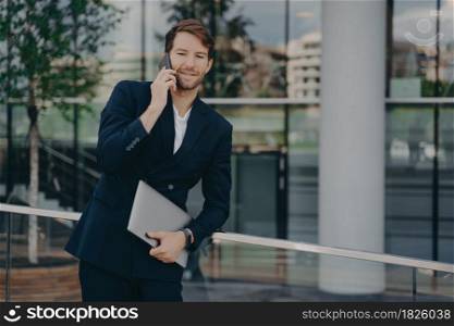 Confident young bearded businessman dressed in formal clothes making business call, standing outside of glass building with laptop in relaxed pose while talking on mobile phone with colleagues. Confident young businessman standing outside with laptop computer and talking on mobile phone