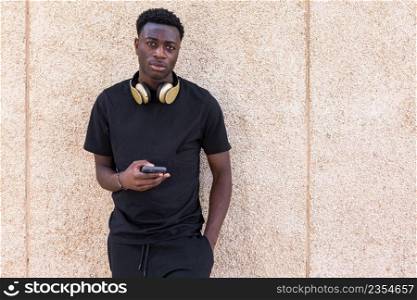 Confident young black man in casual outfit and headphones on neck using smartphone and looking at camera while standing near beige concrete wall with hand in pocket. Stylish young African American male browsing mobile phone near beige wall