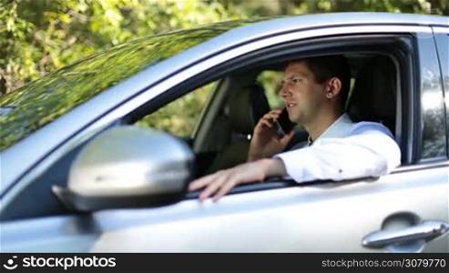 Confident young businessman communicating with business partner on smartphone and gesturing while sitting in luxury car. Handsome businessman arguing with colleague using mobile phone while driving automobile.