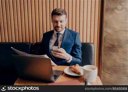 Confident young businessman in formal clothes enjoying coffee and trying to keep up with busy work schedule on his smartphone, working on laptop and reading newspaper while sitting at coffee shop. Confident young businessman in formal clothes enjoying coffee and working remotely in cafe