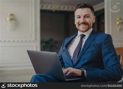 Confident young businessman in formal suit, working with laptop in office, smiling at camera, feeling happy. Business occupation concept.