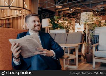 Confident young businessman reading newspaper and latest financial news articles while sitting in armchair in modern cafe shop interior while waiting for work colleague to have morning coffee together. Positive young businessman reading newspaper in cafe