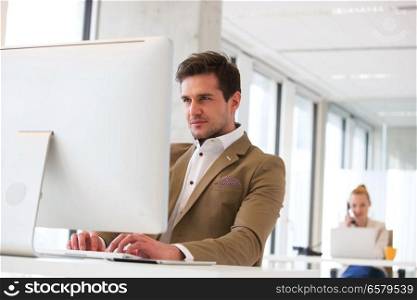 Confident young businessman working on computer with female colleague in background at office