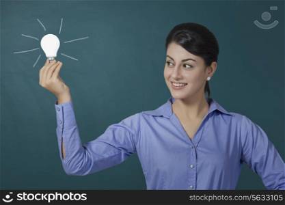 Confident young businesswoman holding light bulb against green board at office