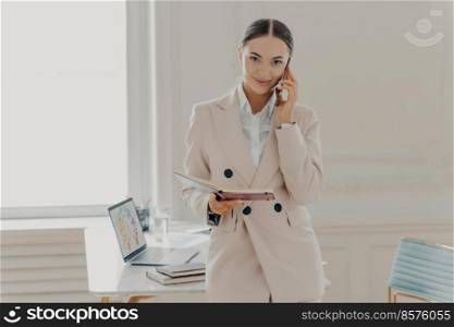 Confident young businesswoman talking via mobile phone, standing in light interior of modern office and calling business partner, smiling female director in beige formal suit discussing business ideas. Young business lady talking on phone and discussing business ideas