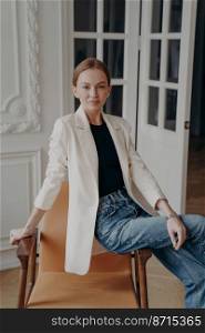 Confident young caucasian woman wearing suit jacket, posing for portrait, sitting on armchair indoors. Pretty stylish businesswoman in casual clothing sits on chair, looking at camera.. Confident pretty young businesswoman wearing casual clothing, sitting on stylish armchair indoors
