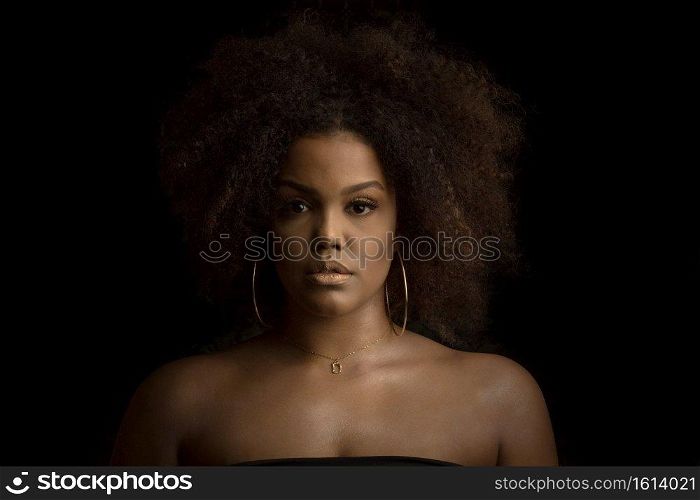 Confident young curly haired African American female with perfect makeup and hoop earrings and necklace looking at camera against black background. Beautiful black woman with makeup and bijouterie