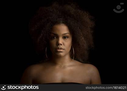 Confident young curly haired African American female with perfect makeup and hoop earrings and necklace looking at camera against black background. Beautiful black woman with makeup and bijouterie