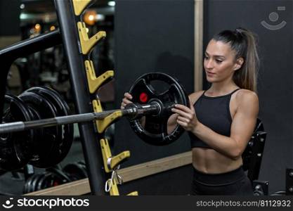 Confident young fit female athlete with long dark hair in sportswear putting weight plate on barbell while working out in modern sports club. Muscular sportswoman putting weight plate on barbell in gym