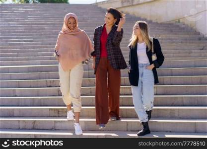 Confident young happy multiracial women students in casual outfits and hijab smiling while walking downstairs in university campus on sunny day. Positive young diverse women friends walking downstairs in campus
