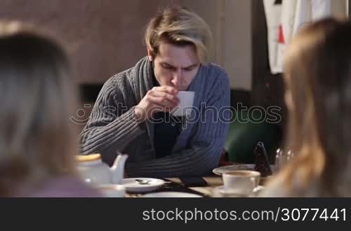 Confident young hipster sitting at cafe table and surfing net on cell phone while spending time together with teenage friends at coffee shop. Teenager drinking coffee and browsing social media with mobile phone during meeting with groupmates in cafe.