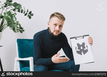 confident young male psychologist sitting arm chair showing rorschach inkblot test paper