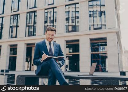 Confident young man dressed formally writing something in his notebook while sitting on bench with modern open laptop with information, working remotely. Fountain behind him in blurred background. Confident young man in smart casual wear writing something in his notebook while sitting on bench