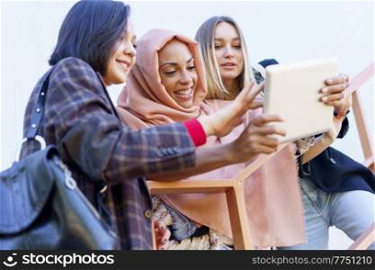 Confident young multiracial female girlfriends smiling and sharing tablet standing on metal staircase on street. Smiling young diverse ladies using tablet on stairs