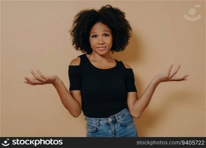 Confused Afro woman, wavy hair, raises hands in bewilderment, can’t decide. Isolated on beige background