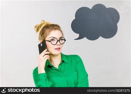 Confused business woman wearing green shirt and red eyeglasses talking on phone with black thinking or speech bubble next to her.. Business woman talking on phone with thinking bubble