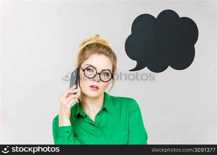 Confused business woman wearing green shirt and red eyeglasses talking on phone with black thinking or speech bubble next to her.. Business woman talking on phone with thinking bubble