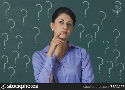 Confused young businesswoman standing against question marks on green board with question marks at office